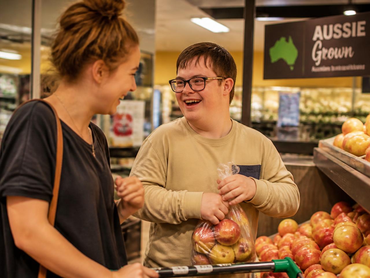 A young adult with Down syndrome shopping for fruit with a friend at a local grocer, both laughing.
